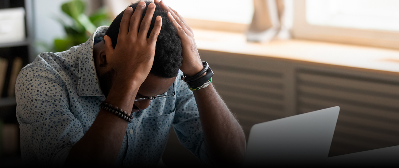 A man sits at his desk, looking stressed with his hands on his head.